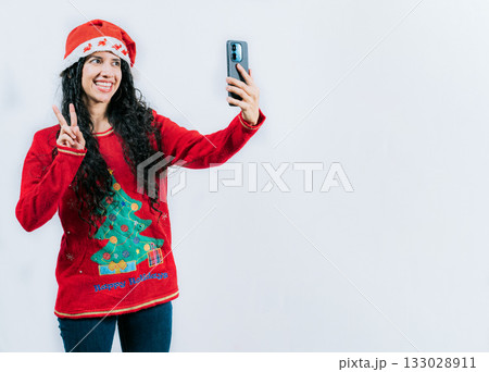 Happy young latin woman in Christmas sweater taking selfies isolated. Funny girl in Christmas hat and sweater taking selfie, isolated 133028911