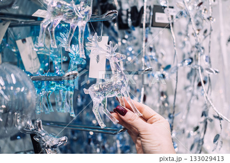 Hand holding a transparent glass reindeer ornament in a store aisle filled with shimmering Christmas decorations. Sustainable holiday shopping, eco-friendly materials, mindful gifting Hand holding a transparent glass reindeer ornament in a store aisle filled with shimmering Christmas decorations. Sustainable holiday shopping, eco-friendly materials, mindful gifting 133029413