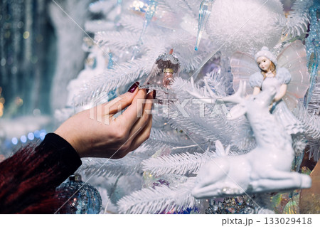 Hand placing a delicate bell ornament on a white Christmas tree adorned with fairy and reindeer decorations. Holiday bonding moments, personal touch in decor, festive rituals, seasonal creativity 133029418