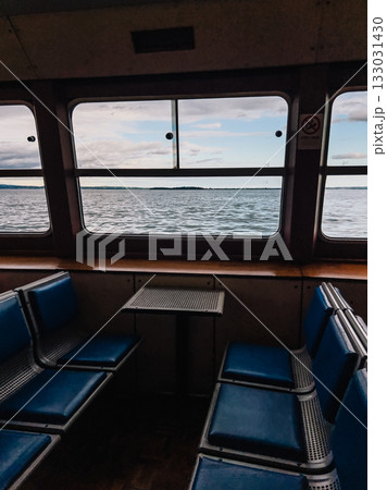 Empty passenger seats and small table inside ferry boat navigating on Lake with view on water and cloudy sky through large windows. Vacant ferry seats, small table, large windows revealing Lake waters Empty passenger seats and small table inside ferry boat navigating on Lake with view on water and cloudy sky through large windows. Vacant ferry seats, small table, large windows revealing Lake waters 133031430