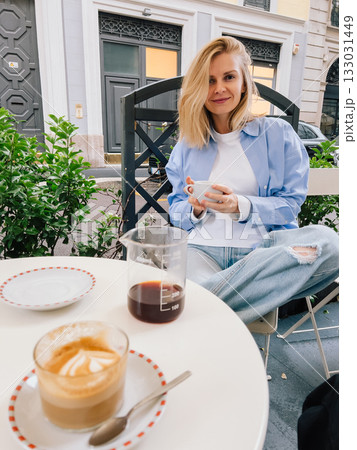 Young woman sitting at table in bustling street cafe, savoring rich cup of coffee and creamy cappuccino while dressed in casual attire and enjoying vibrant urban atmosphere. Woman sipping cappuccino Young woman sitting at table in bustling street cafe, savoring rich cup of coffee and creamy cappuccino while dressed in casual attire and enjoying vibrant urban atmosphere. Woman sipping cappuccino 133031449