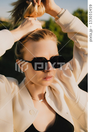 Blonde woman with sunglasses is tying her hair into ponytail outdoors, enjoying warm sunlight of golden hour, wearing white jacket and black top. Portrait of blonde woman securing hair in sunglasses Blonde woman with sunglasses is tying her hair into ponytail outdoors, enjoying warm sunlight of golden hour, wearing white jacket and black top. Portrait of blonde woman securing hair in sunglasses 133031459