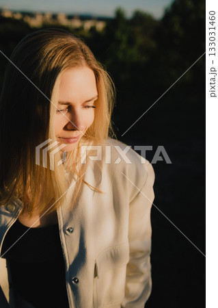 Young blonde woman wearing light jacket is smiling and looking down, enjoying warm sunlight during golden hour, with blurred background of trees and cityscape. Woman basking in golden hour sunlight Young blonde woman wearing light jacket is smiling and looking down, enjoying warm sunlight during golden hour, with blurred background of trees and cityscape. Woman basking in golden hour sunlight 133031460