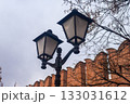 Vintage black forged street lamp against an old red brick fortress wall with battlements. Urban background for the concept of history, security, and city street illumination. 133031612