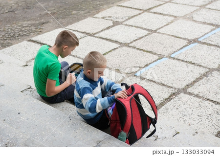 Two boys getting ready on stone steps before school. One packs a red backpack, the other uses an electronic device in his black bag. Two boys getting ready on stone steps before school. One packs a red backpack, the other uses an electronic device in his black bag. 133033094