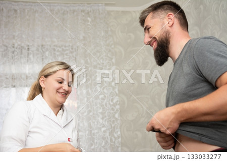 Bearded man smiling at female healthcare provider, possibly receiving medical consultation or checkup, in a room with patterned wallpaper. Bearded man smiling at female healthcare provider, possibly receiving medical consultation or checkup, in a room with patterned wallpaper. 133033277