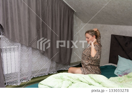 Woman in animal print robe sits on her unmade bed pulling her blonde hair up into a ponytail in a minimally decorated bedroom. 133033278