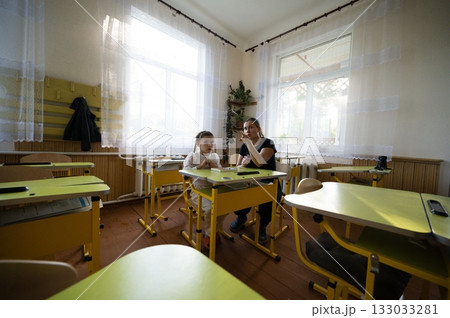 Teacher and young student in classroom. The pair sit at desks under bright windows in the well lit educational space. Teacher and young student in classroom. The pair sit at desks under bright windows in the well lit educational space. 133033281