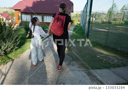 A mother and daughter walking away hand in hand on a path toward buildings in the background on a bright sunny day outside. A mother and daughter walking away hand in hand on a path toward buildings in the background on a bright sunny day outside. 133033284