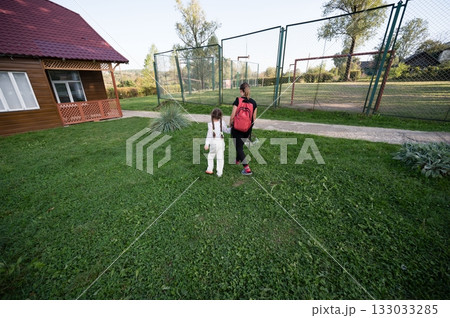 Mother and daughter stroll along a grassy path near a cozy cabin and fenced-in field, enjoying a peaceful day outdoors together. 133033285
