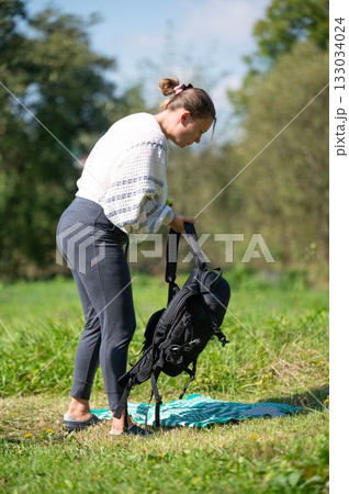 Woman prepares for a picnic in a grassy field, setting out a blanket and backpack in the sunny outdoors, looking down at the scene. Woman prepares for a picnic in a grassy field, setting out a blanket and backpack in the sunny outdoors, looking down at the scene. 133034024