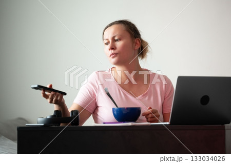 Woman working from home, holding phone while eating. A laptop is next to a bowl of food on a dark desk during a casual day indoors. 133034026