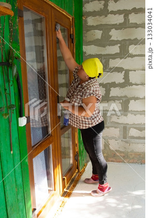Woman cleaning glass door on the patio of her house, with cleaning product and a rag, during the day, wearing a yellow kerchief. 133034041