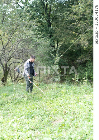 Man trimming tall grass in a forest clearing using a string trimmer to cut back overgrowth. Summer landscaping maintenance in progress. 133034176