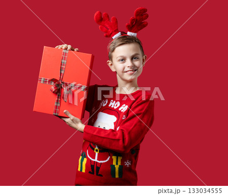 Cheerful boy in festive Santa sweater and reindeer antlers holding red Christmas gift box with bow, standing on red background 133034555