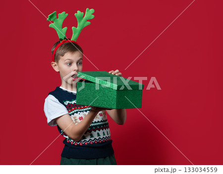 Happy boy with green antlers holds stack of red and green Christmas gift boxes, smiling in front of red background in festive clothes Happy boy with green antlers holds stack of red and green Christmas gift boxes, smiling in front of red background in festive clothes 133034559