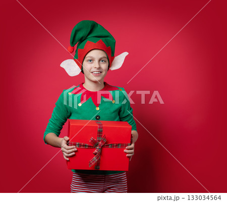 Merry Christmas concept. Smiling funny boy in Santa's elf costume holding Christmas gift in his hand. Shooting on red background. 133034564