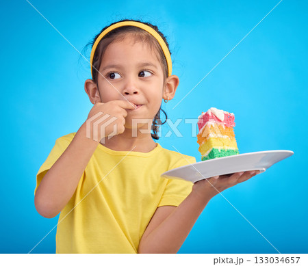 Birthday cake, taste and young girl with a sweet in studio eating icing for party event dessert. Celebration, rainbow food and hungry kid in a isolated and blue background studio ready to celebrate 133034657