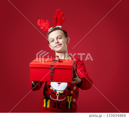 Cheerful boy in festive Santa sweater and reindeer antlers holding red Christmas gift box with bow, standing on red background Cheerful boy in festive Santa sweater and reindeer antlers holding red Christmas gift box with bow, standing on red background 133034869