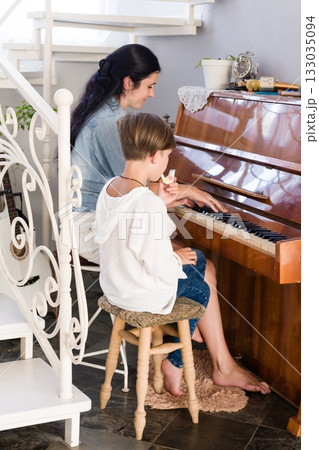 A woman teaches a child to play the piano. They sit together at a vintage instrument in a home setting, focusing on the music. A woman teaches a child to play the piano. They sit together at a vintage instrument in a home setting, focusing on the music. 133035094