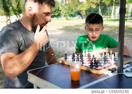 Father and son enjoy a sunny afternoon playing chess in the park, a moment of focused concentration and playful competition. 133035230