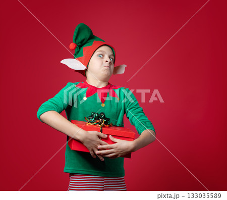 Happy boy dressed as surprise , impression, emotions Christmas elf holding a red present with ribbon, green red costume with pointy hat and elf ears 133035589