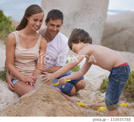 Family, child and building sand castle at beach in summer for fun, travel or holiday with love. A man, woman and kid playing together on vacation at sea with a toy bucket, development and happiness 133035788