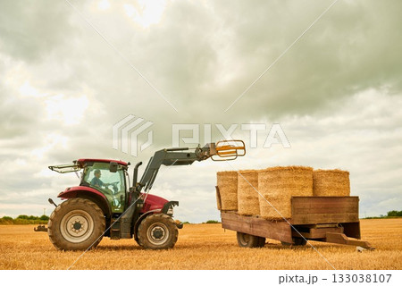 Hay, countryside and a tractor on a farm for sustainability outdoor on an open field during the harvest season. Agriculture, sky and clouds with a red industrial farming vehicle harvesting in nature Hay, countryside and a tractor on a farm for sustainability outdoor on an open field during the harvest season. Agriculture, sky and clouds with a red industrial farming vehicle harvesting in nature 133038107