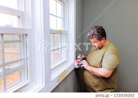 Man sanding wood trim near window during home renovation in bright room 133039385