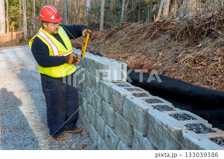 Construction worker measures stone blocks  wall building along gravel pathway during work day hours 133039386