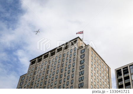 Tall office building with national flag on rooftop and airplane flying in cloudy blue sky Tall office building with national flag on rooftop and airplane flying in cloudy blue sky 133041721