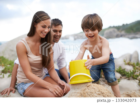 Family, child and sand castle at beach in summer for fun, travel or holiday with a smile. A man, woman and excited kid playing together on vacation at sea with a toy bucket, development and happiness 133042073