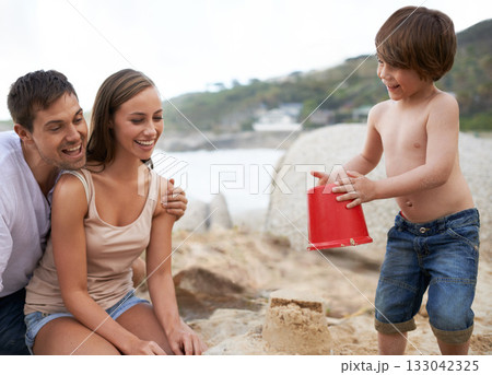 Child, family and sand castle at beach in summer for fun, travel or holiday with a smile. A man, woman and excited kid playing together on vacation at sea with a toy bucket, development and happiness 133042325