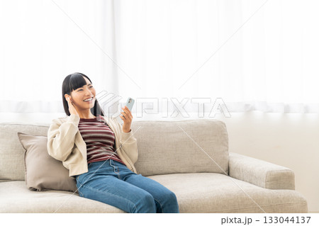 ソファーに座ってスマホで音楽を聴いている若い女性 133044137