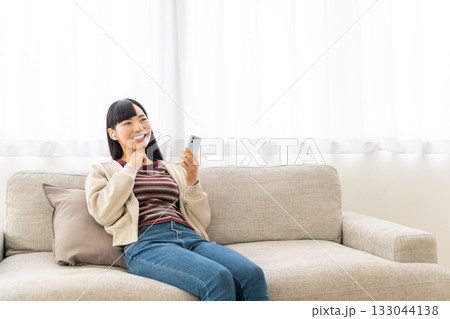 ソファーに座ってスマホで音楽を聴いている若い女性 133044138