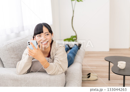 ソファーに寝そべってスマホで音楽を聴いている若い女性 ソファーに寝そべってスマホで音楽を聴いている若い女性 133044263