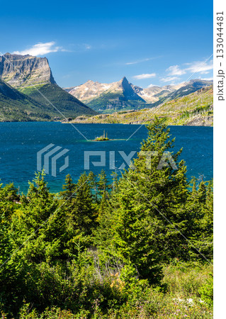 Vertical view of Wild Goose Island in the deep blue Saint Mary Lake, Glacier National Park. The Rocky Mountains rise in the background, framed by pine trees under a blue sky Vertical view of Wild Goose Island in the deep blue Saint Mary Lake, Glacier National Park. The Rocky Mountains rise in the background, framed by pine trees under a blue sky 133044481