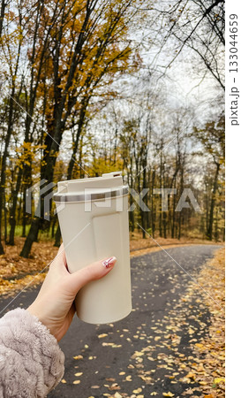 Woman's hand holding a beige travel mug while walking on an autumn park path covered with leaves 133044659