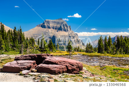 Scenic view of Mount Oberlin from Logan Pass in Glacier National Park. Red argillite rocks are in the foreground, next to an alpine stream and pine trees, under a clear blue sky 133046568