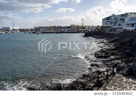 A scenic view of a coastal town in Lanzarote, showcasing the marina, boats, and the rugged shoreline under a cloudy sky. 133046982