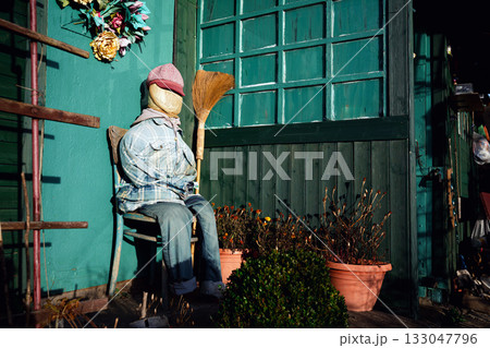 A scarecrow sitting on a wooden chair in front of a small garden gazebo on an allotment, cleverly imitating a resting human figure, basking peacefully in the warm sunlight of a golden autumn day. 133047796
