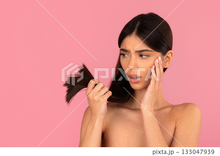Young woman with long hair examining dry hair ends with worried expression 133047939