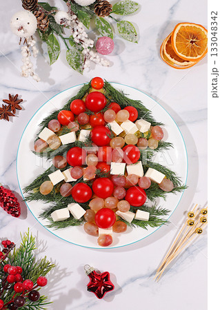 Traditional holiday salad in the form of a live edible Christmas tree with feta cheese, olives, tomatoes and grapes, Christmas and New Year's dishes, food design idea, fir branches and decorations Traditional holiday salad in the form of a live edible Christmas tree with feta cheese, olives, tomatoes and grapes, Christmas and New Year's dishes, food design idea, fir branches and decorations 133048342