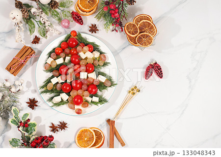 Traditional holiday salad in the form of a live edible Christmas tree with feta cheese, olives, tomatoes and grapes, Christmas and New Year's dishes, food design idea, fir branches and decorations 133048343