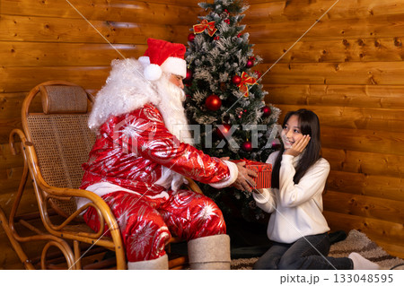 Girl is receiving a gift box from Santa in front of a decorated Christmas tree. 133048595