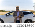 man is standing in front of a white car, looking down at an object in his hand 133048690