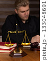 The judge is sitting at a desk with a balance scale, books, and a gavel 133048691