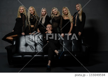 Group of six women in black outfits with a man posing on a black sofa in a studio setting 133049594