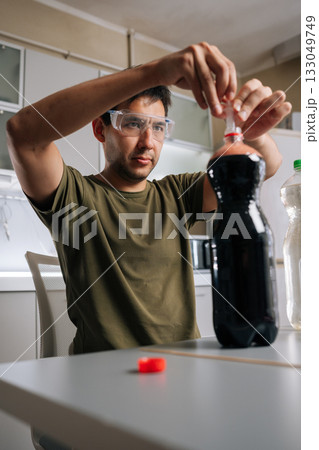 Vertical portrait of scientist wearing safety glasses preparing fertilizer using syringe and soda bottle filled with dark liquid, surrounded by various lab equipment in makeshift kitchen laboratory. 133049749