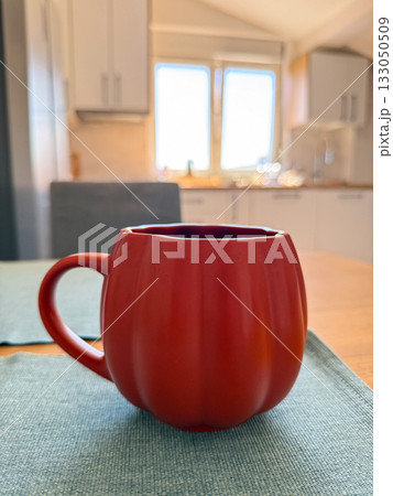 Red ceramic mug on kitchen table with soft sunlight. Morning warmth, comfort and everyday home ritual. 133050509